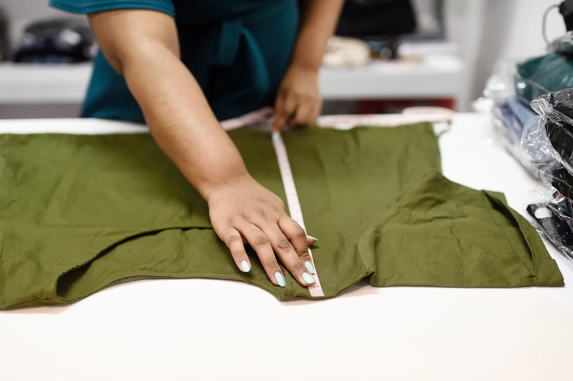 Rani's hands running a measuring tape along an olive-green top laid flat on a table