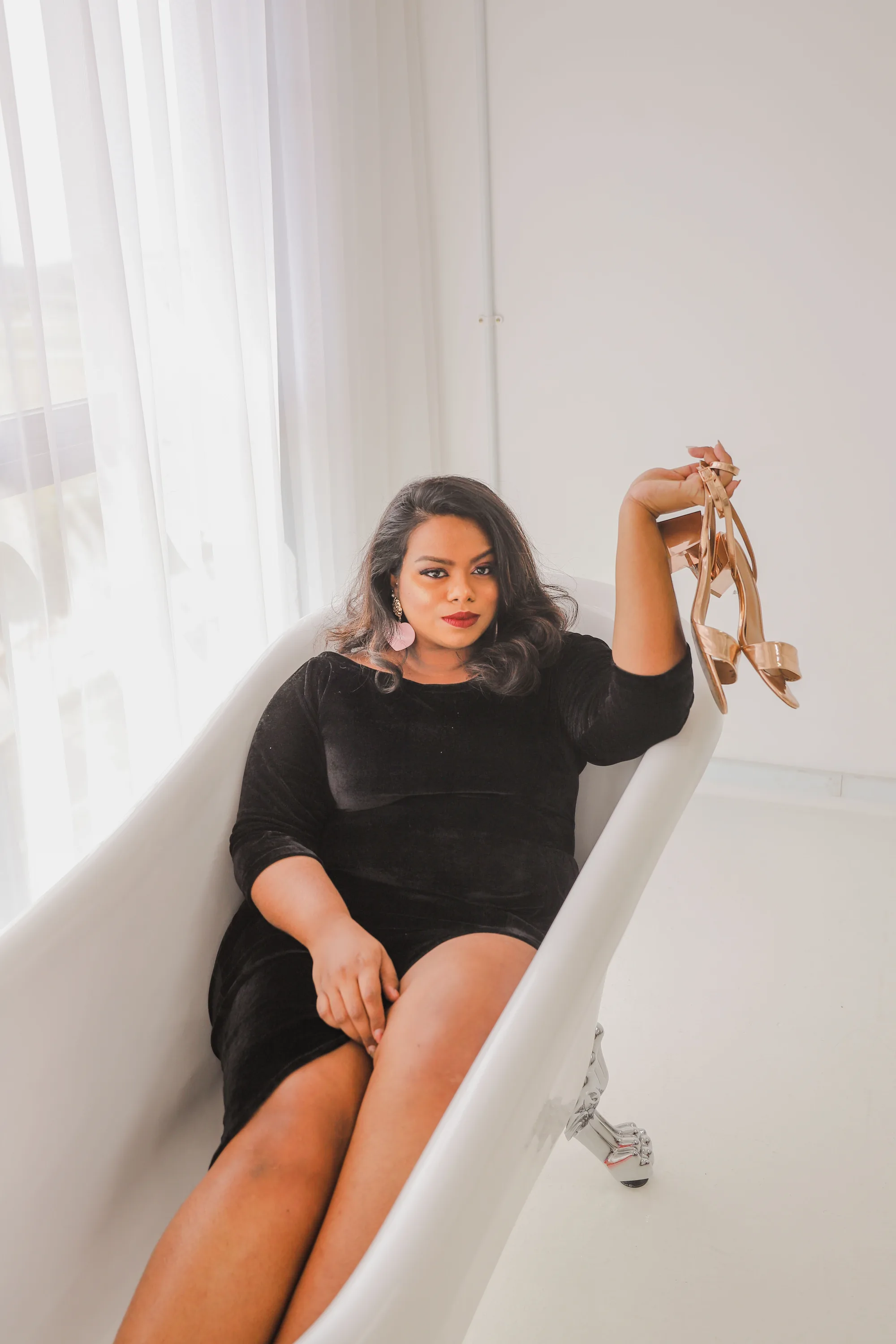 Rani lounging in a white bathtub in a black velvet dress, holding a pair of heels up in one hand