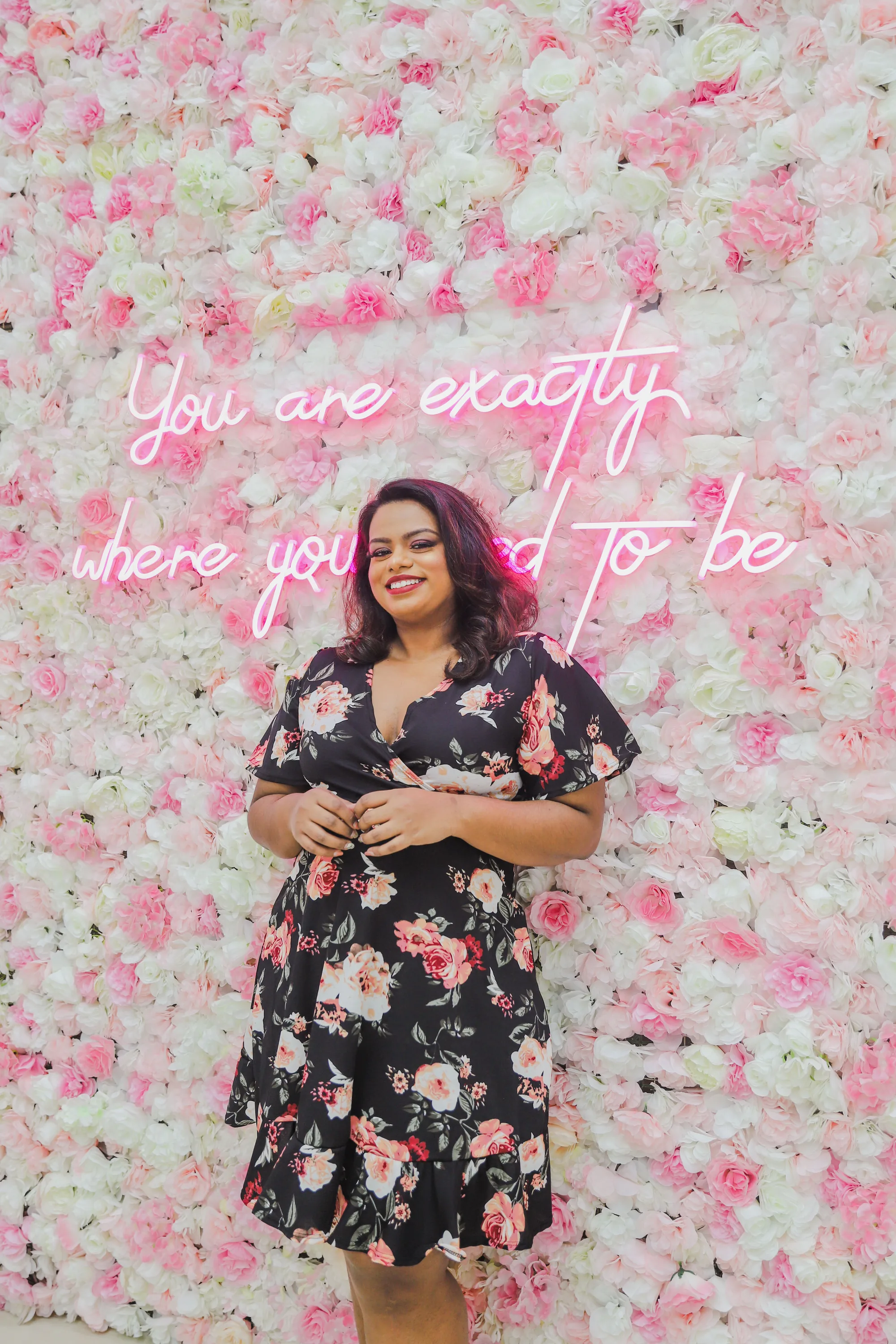 Rani smiling in a floral dress in front of a wall of pink and white flowers with a neon sign reading 'You are exactly where you need to be'
