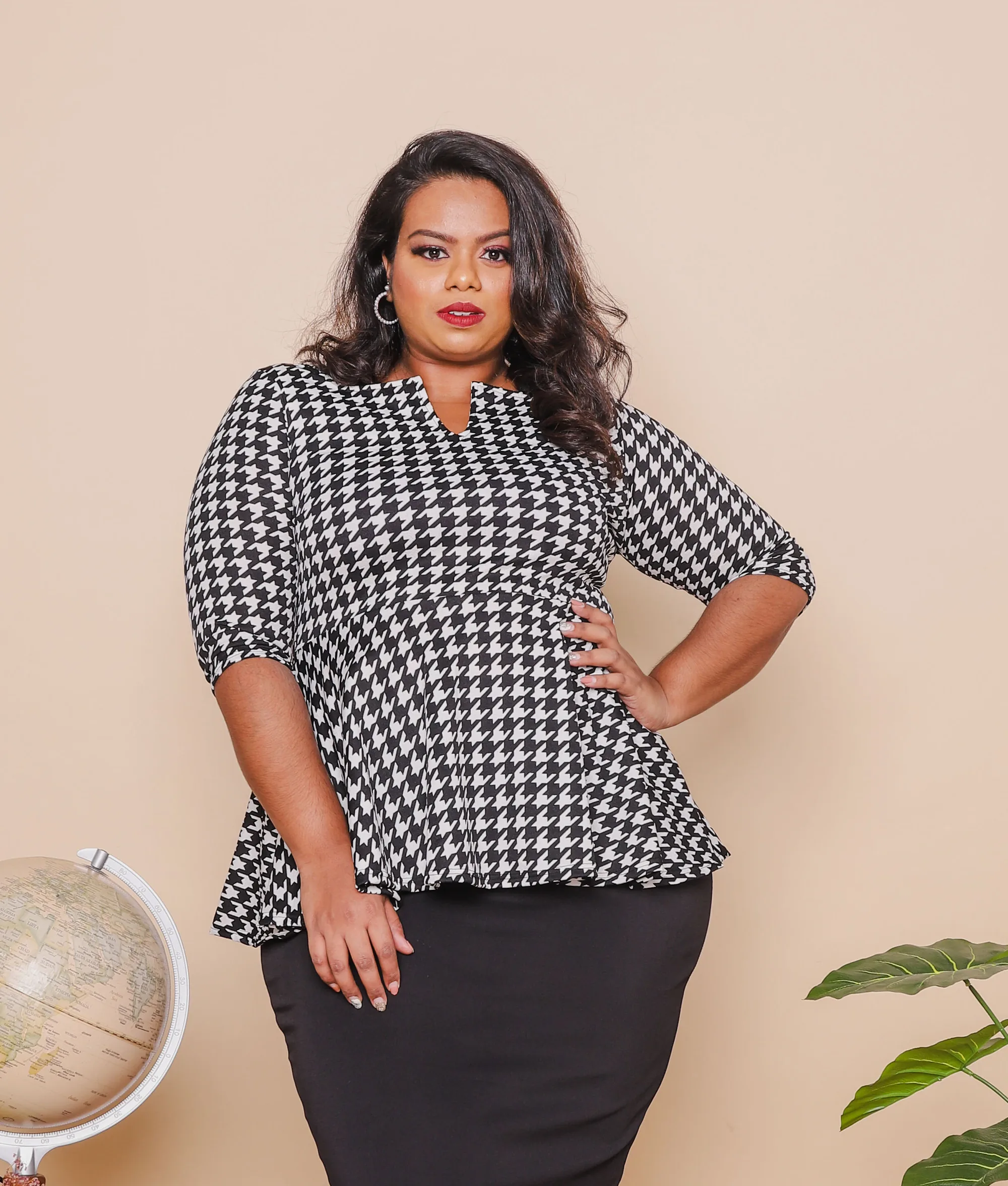 Rani standing with a hand on her hip in a black-and-white houndstooth peplum top and black pencil skirt, beside a globe and a potted plant