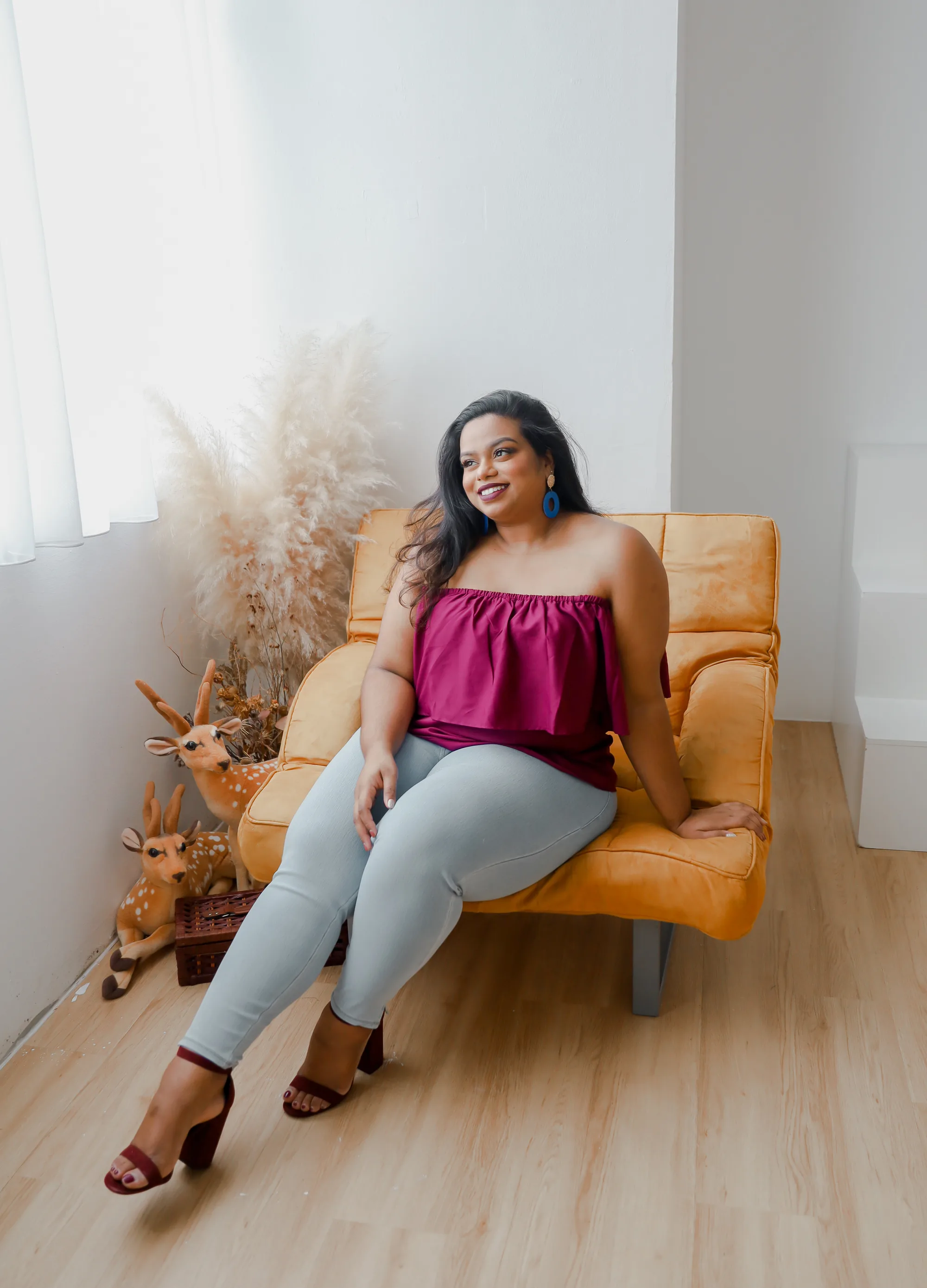 Rani relaxed on a mustard-yellow sofa in a plum off-the-shoulder top and pale jeans, smiling, beside dried pampas grass