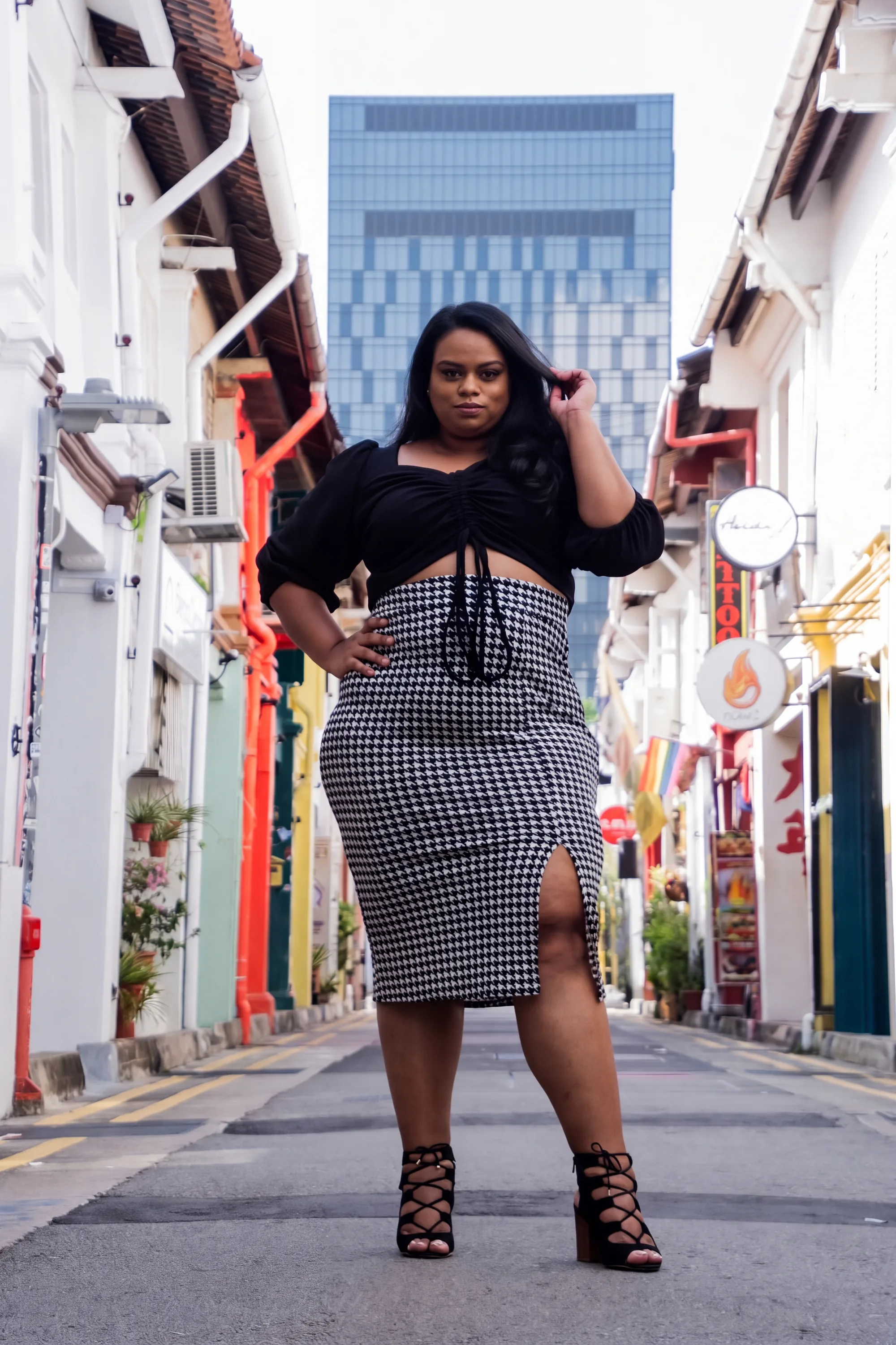 Rani standing in the middle of a street of colourful shophouses, hand in her hair, in a black ruched crop top and a houndstooth pencil skirt with a front split