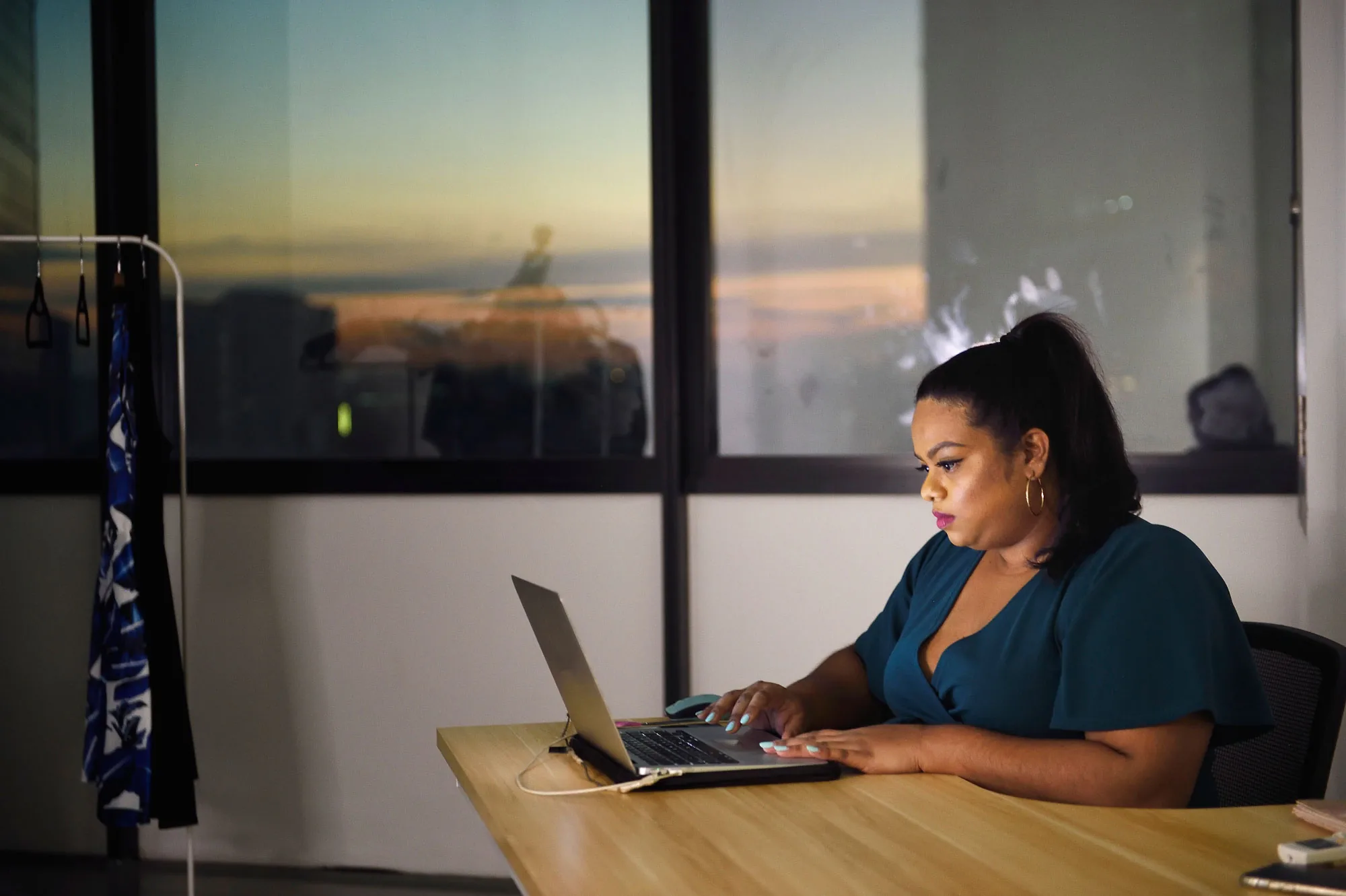 Rani working on a laptop at a wooden desk beside a rail of clothes, city sunset in the window