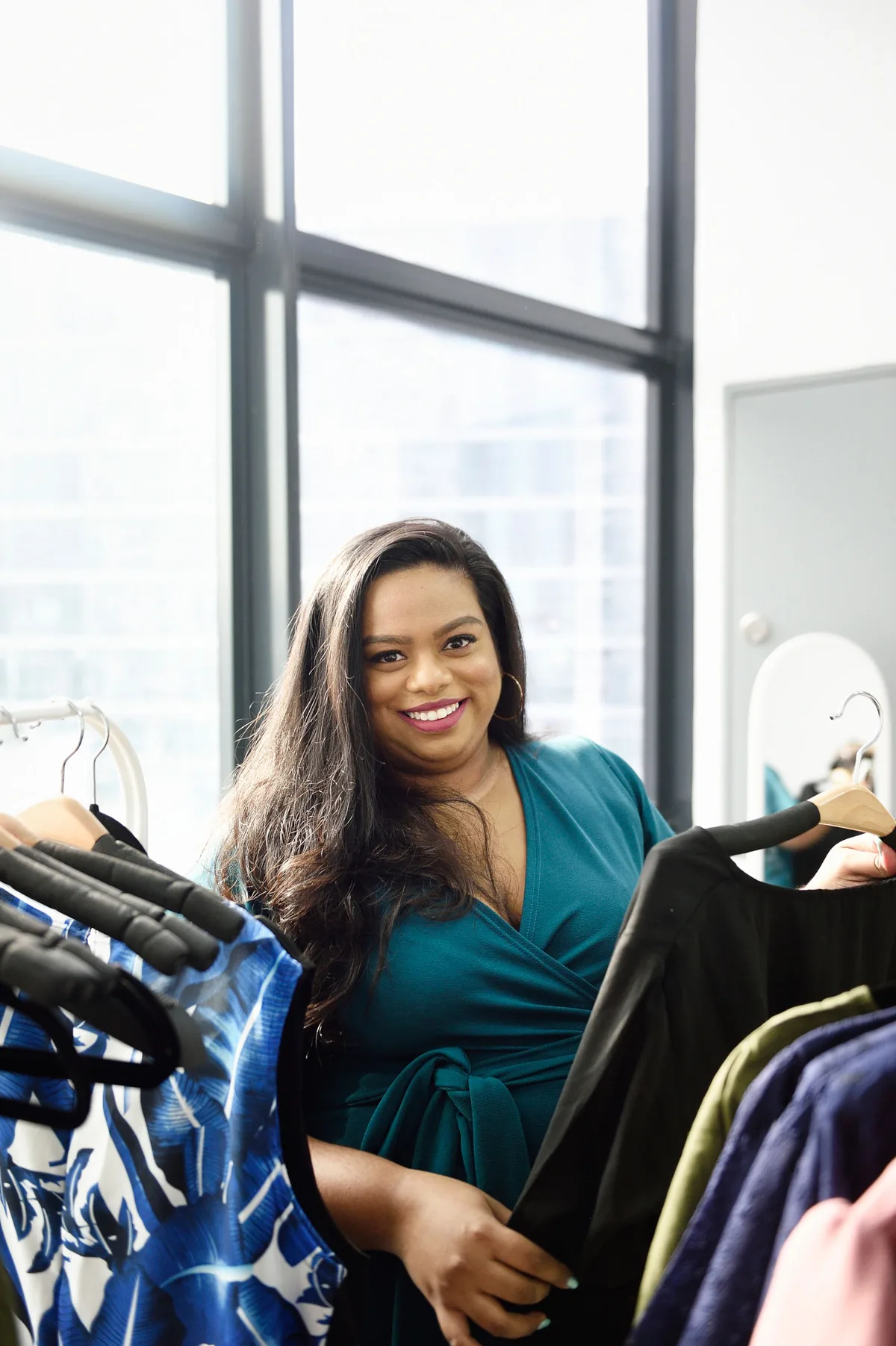 Rani smiling in a teal wrap dress as she looks through a rail of clothes by a tall window